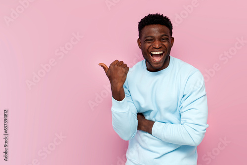 african american man in blue sweater laughs and points his hand to the side on pink isolated background