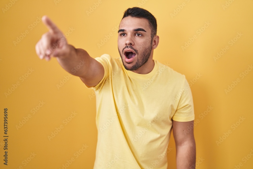 Young hispanic man standing over yellow background pointing with finger surprised ahead, open mouth amazed expression, something on the front Young hispanic man standing over yellow background pointing with finger surprised ahead, open mouth amazed expression, something on the front
