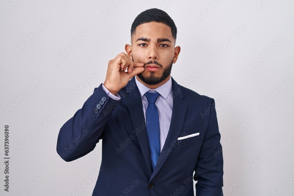 Young hispanic man wearing business suit and tie mouth and lips shut as zip with fingers. secret and silent, taboo talking Young hispanic man wearing business suit and tie mouth and lips shut as zip with fingers. secret and silent, taboo talking