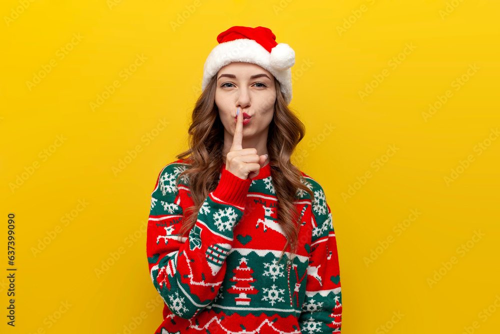 girl in christmas sweater and santa claus hat holds finger on her lips and shows gesture of silence on yellow background girl in christmas sweater and santa claus hat holds finger on her lips and shows gesture of silence on yellow background