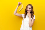 young girl holding small banana and mocking on yellow isolated background, woman presents erotic and intimate concept