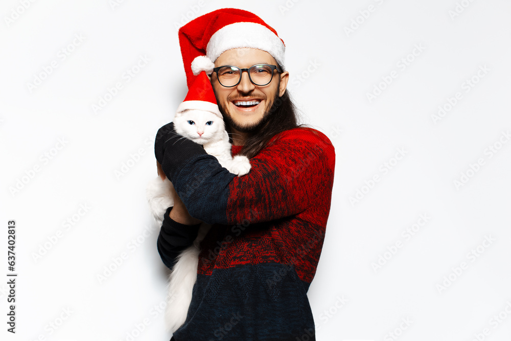 Studio portrait of young happiness man with eyeglasses in red sweater hugging a white kitty cat with blue eyes, both wearing Santa Claus hat. Isolated on white background.