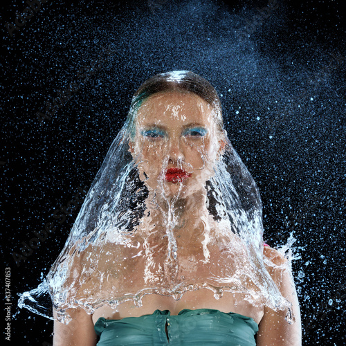 Portrait of wet young girl with bare shoulders with bright and colorful makeup on face and stream of water isolated dark mode background.