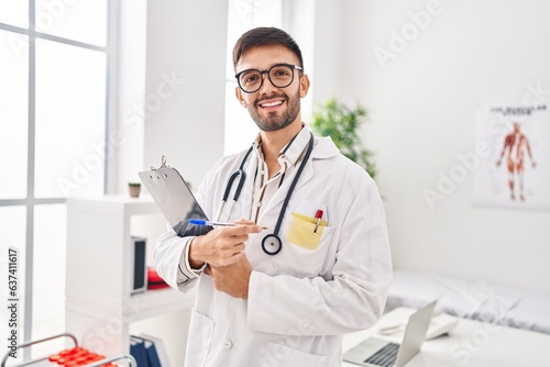 Young hispanic man doctor smiling confident holding clipboard at clinic