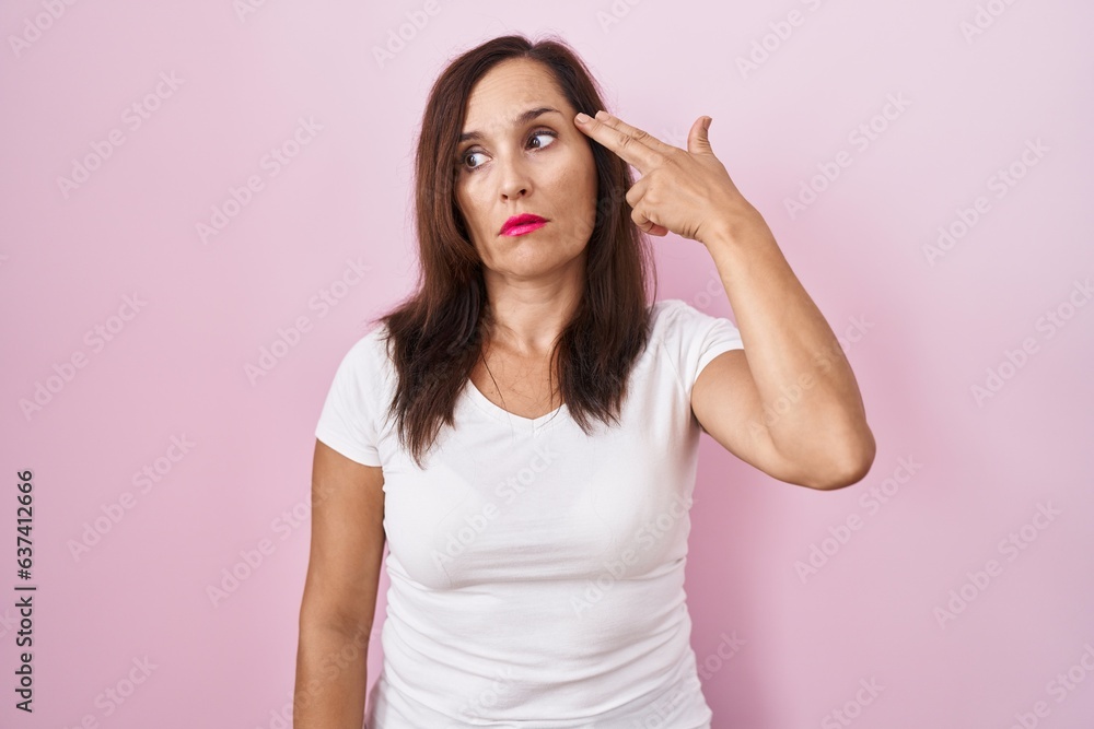 Middle age brunette woman standing over pink background shooting and killing oneself pointing hand and fingers to head like gun, suicide gesture. Middle age brunette woman standing over pink background shooting and killing oneself pointing hand and fingers to head like gun, suicide gesture.