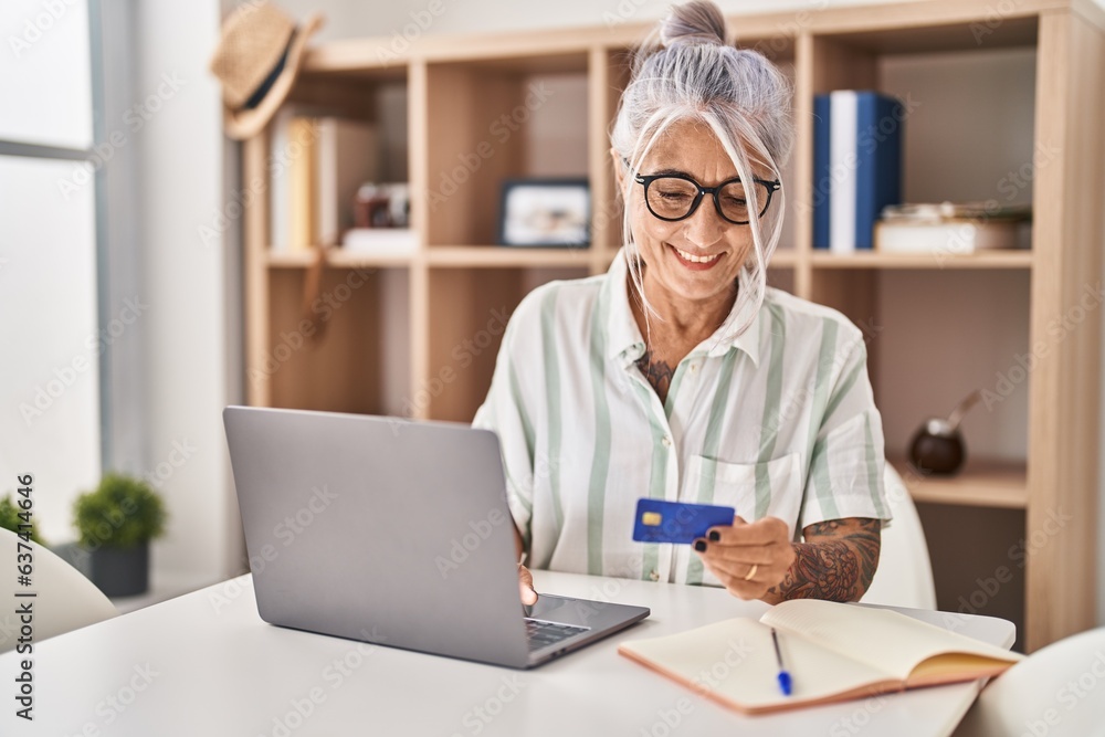 Middle age grey-haired woman using laptop and credit card sitting on table at home Middle age grey-haired woman using laptop and credit card sitting on table at home