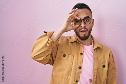 Young hispanic man standing over pink background doing ok gesture shocked with surprised face, eye looking through fingers. unbelieving expression.