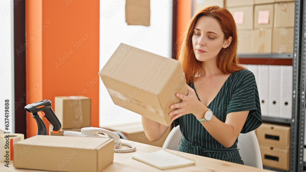 Young redhead woman ecommerce business worker holding package sitting on table at office Young redhead woman ecommerce business worker holding package sitting on table at office