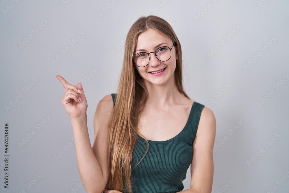 Young caucasian woman standing over white background with a big smile on face, pointing with hand finger to the side looking at the camera.