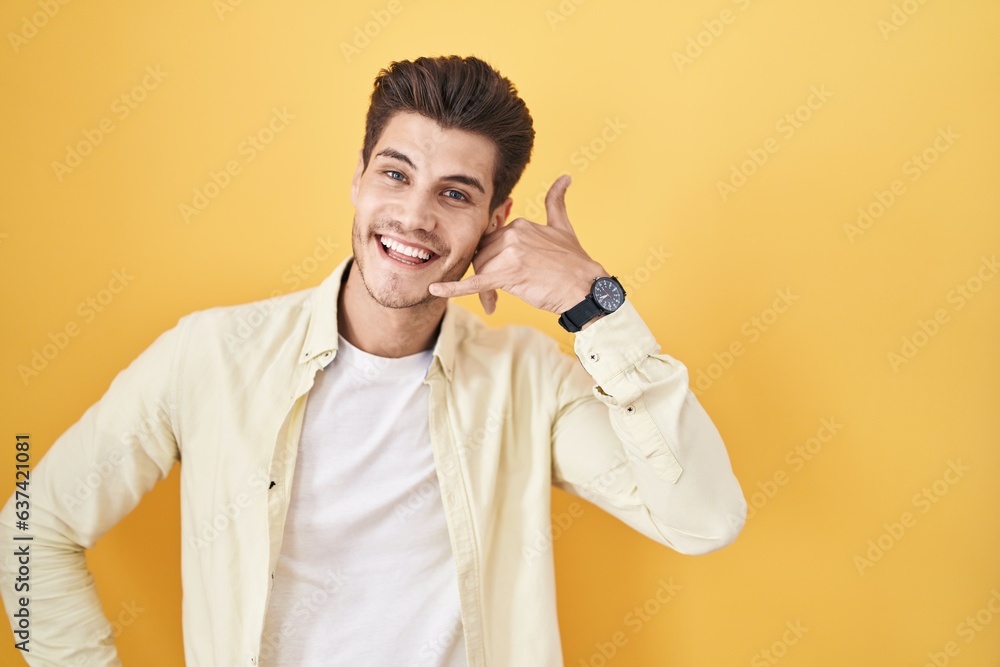 Young hispanic man standing over yellow background smiling doing phone gesture with hand and fingers like talking on the telephone. communicating concepts. Young hispanic man standing over yellow background smiling doing phone gesture with hand and fingers like talking on the telephone. communicating concepts.