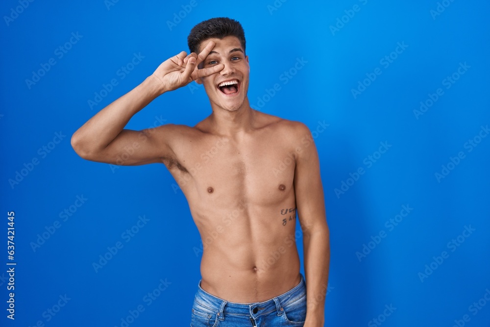 Fototapeta premium Young hispanic man standing shirtless over blue background doing peace symbol with fingers over face, smiling cheerful showing victory