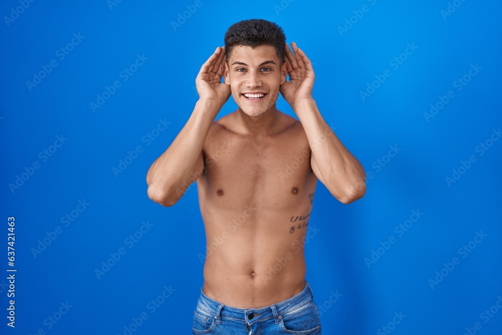 Fototapeta premium Young hispanic man standing shirtless over blue background trying to hear both hands on ear gesture, curious for gossip. hearing problem, deaf