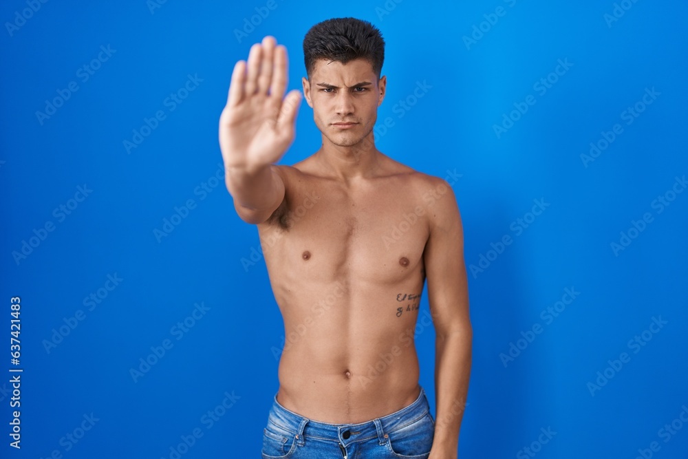 Young hispanic man standing shirtless over blue background doing stop sing with palm of the hand. warning expression with negative and serious gesture on the face.