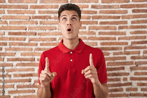 Young hispanic man standing over bricks wall amazed and surprised looking up and pointing with fingers and raised arms.