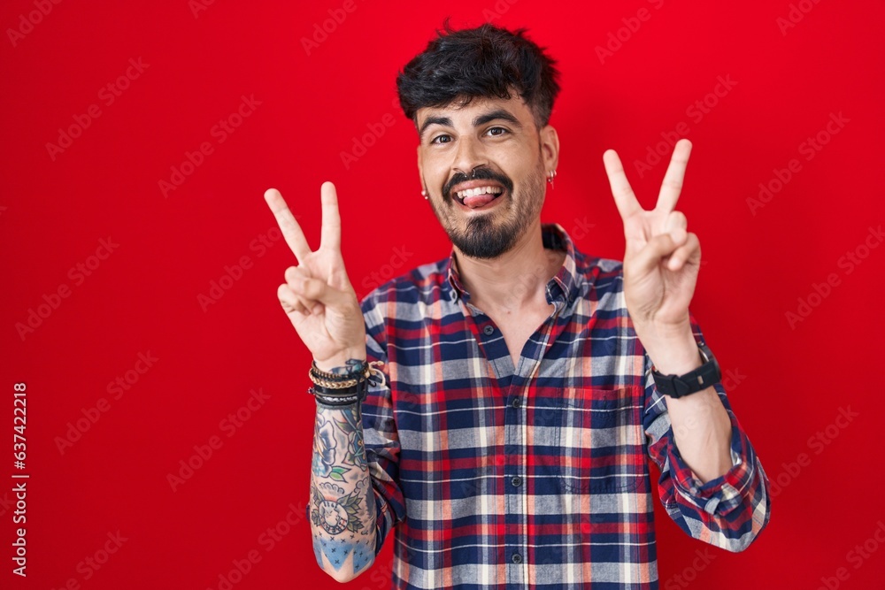 Young hispanic man with beard standing over red background smiling with tongue out showing fingers of both hands doing victory sign. number two. Young hispanic man with beard standing over red background smiling with tongue out showing fingers of both hands doing victory sign. number two.