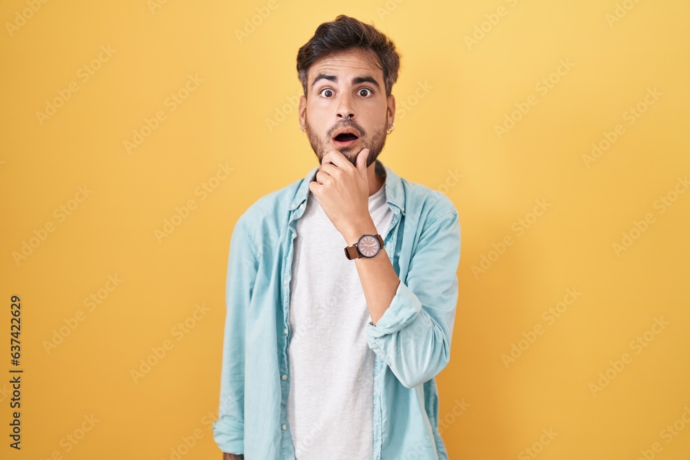 Young hispanic man with tattoos standing over yellow background looking fascinated with disbelief, surprise and amazed expression with hands on chin