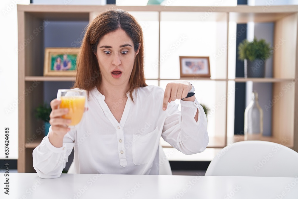 Brunette woman drinking glass of orange juice pointing down with fingers showing advertisement, surprised face and open mouth Brunette woman drinking glass of orange juice pointing down with fingers showing advertisement, surprised face and open mouth