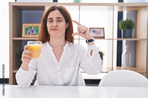Brunette woman drinking glass of orange juice smiling and confident gesturing with hand doing small size sign with fingers looking and the camera. measure concept.