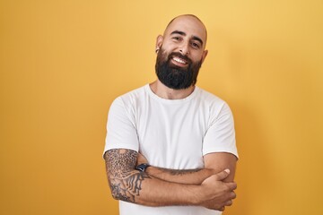 Young hispanic man with beard and tattoos standing over yellow background happy face smiling with crossed arms looking at the camera. positive person.