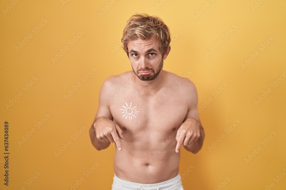 Caucasian man standing shirtless wearing sun screen pointing down looking sad and upset, indicating direction with fingers, unhappy and depressed.
