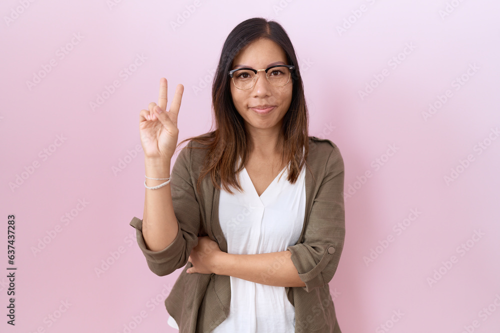 Middle age chinese woman wearing glasses over pink background smiling with happy face winking at the camera doing victory sign. number two. Middle age chinese woman wearing glasses over pink background smiling with happy face winking at the camera doing victory sign. number two.