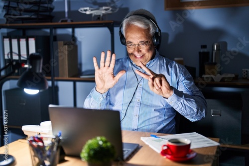 Hispanic senior man wearing call center agent headset at night showing and pointing up with fingers number seven while smiling confident and happy.