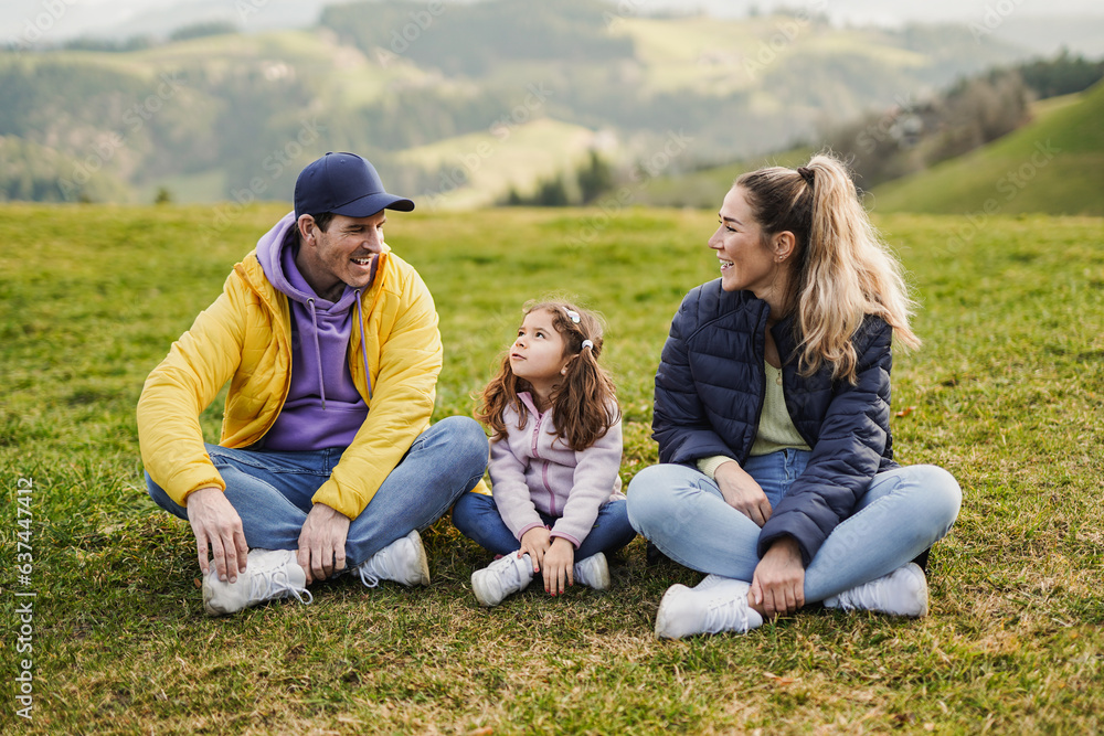Happy family having fun taking a break sitting on grass during hiking day with mountain in background - Active parents with child outdoor Happy family having fun taking a break sitting on grass during hiking day with mountain in background - Active parents with child outdoor