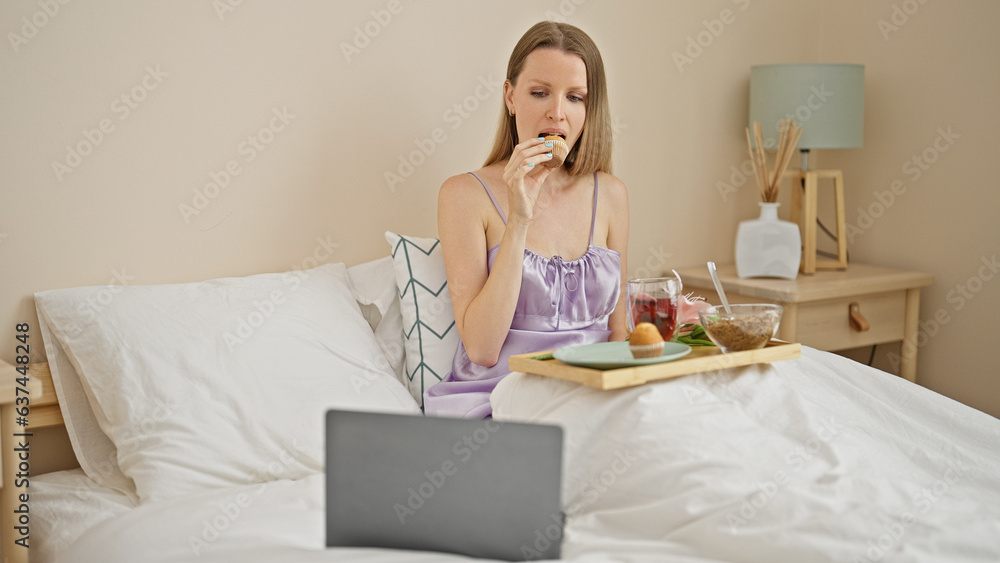 Young blonde woman having gift breakfast watching movie on laptop at bedroom