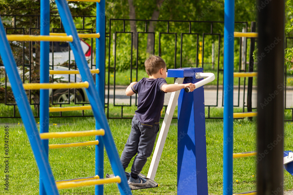 Fototapeta premium A little caucasian athletic boy works out on a street simulator on the playground. A sporty child does exercises in kindergarten. Sports and activities. Healthy lifestyle. Sunny summer day