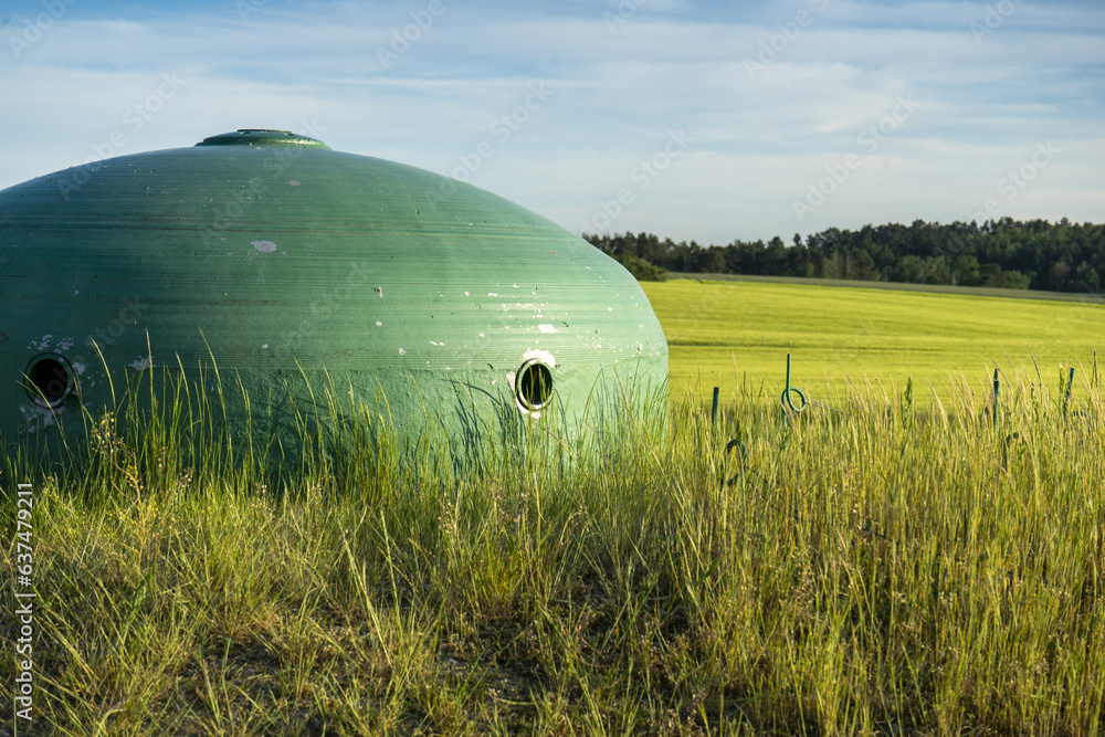 Fototapeta premium Stalowa kopuła bunkra pomalowana na zielono wpasowująca się idealnie w rolniczy krajobraz okolicy