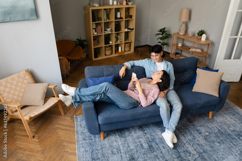 Happy Korean Young Couple Using Phone Relaxing In Living Room