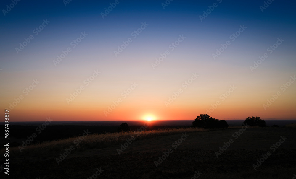 Horizon line with clear blue sky and the sun setting over the horizon with orange tones.