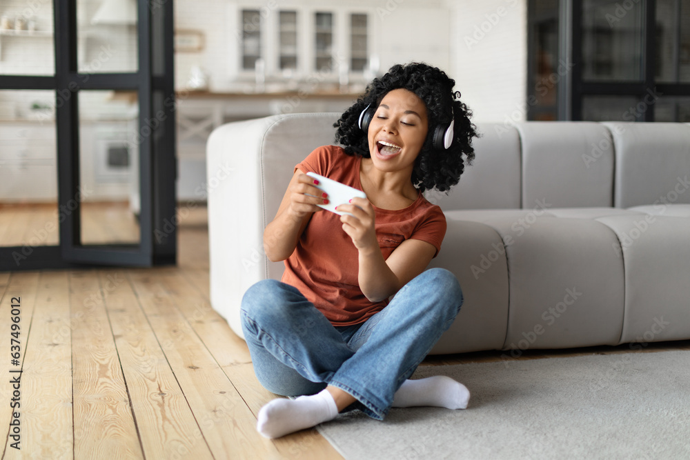 Cheerful Black Woman With Smartphone And Headphones Playing Video Games At Home