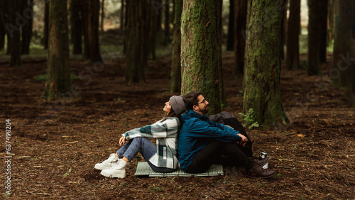 Tired cheerful young caucasian couple in jackets sits in forest on mat, enjoy travel at weekend, rest, relax