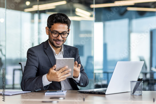 Valokuvatapetti Young successful hispanic businessman at workplace using app on tablet computer, smiling man inside office building, working with laptop, wearing business suit, happy with achievement