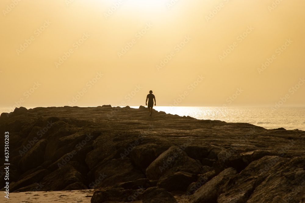 Silhouette of a man wearing wireless headphones walking on a cliff at sunset