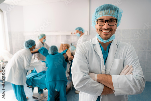 Portrait of a handsome smiling successful medical professional wearing eyeglasses and surgical cap with arms crossed, standing in delivery room in front of medical team and woman in labor.