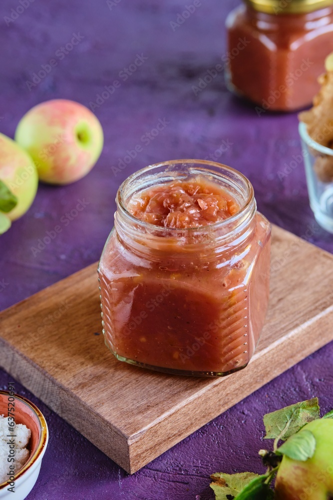 Spicy sauce, apple chutney in a glass jar on a wooden board on a purple concrete background. Indian food. Spicy sauce, apple chutney in a glass jar on a wooden board on a purple concrete background. Indian food.