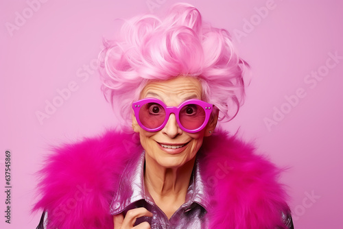 Beautiful elderly fashionable woman in stylish pink clothes, with pink hair and glasses posing on a pink background. Woman looking at the camera in surprise