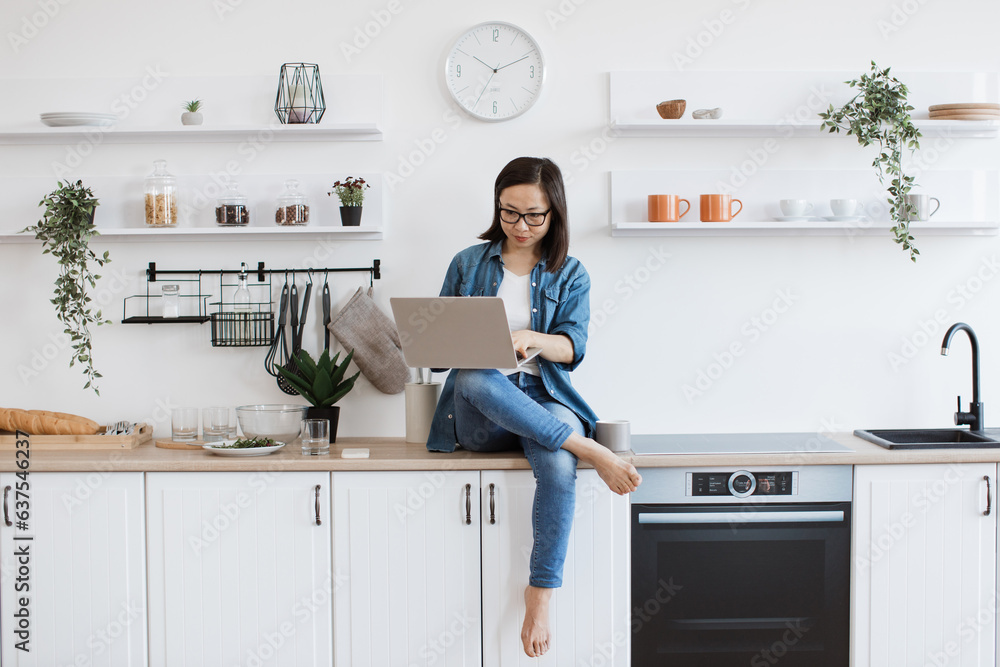 Asian student with computer sitting on worktop in kitchen Asian student with computer sitting on worktop in kitchen