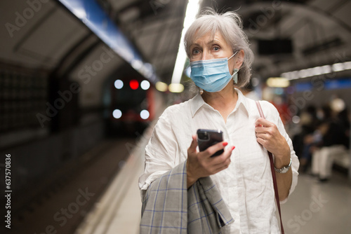 Elderly Caucasian woman in protective mask standing in metro station and using her smartphone while waiting for train arrival.