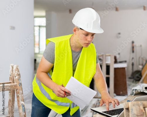 Obraz na plátně Teenage boy wearing yellow vest and hardhat using laptop and checking documentation in construction site
