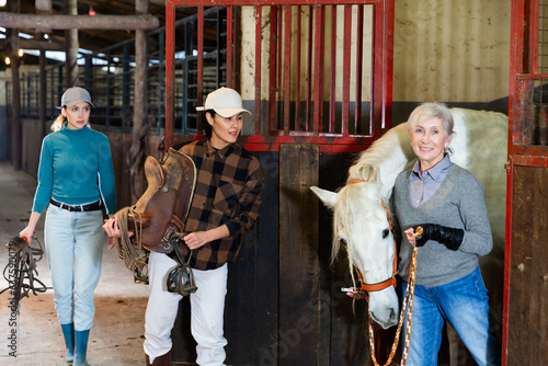 Asian woman stable worker ready to saddle white racehorse being led out of stall by confident older woman rancher..