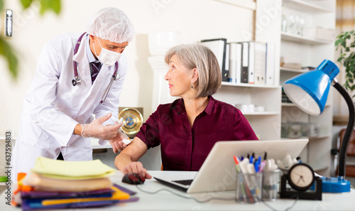 Male doctor in white gown and face mask vaccinating senior woman office worker who sitting at table.