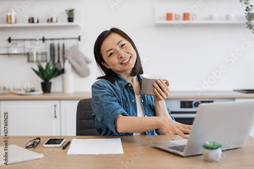 Adult posing with drink while working on laptop in kitchen