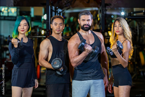 Group portrait of diversity muscular gym trainers standing together while lifting dumbbell inside gym with dark tone background for exercise and workout