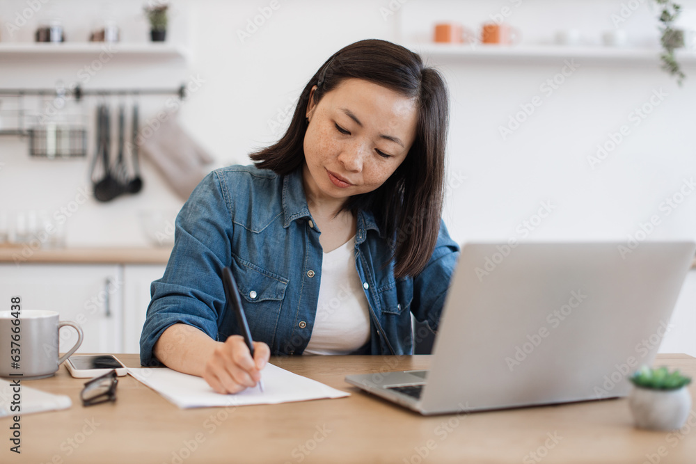 Woman making notes of article from computer in apartment Woman making notes of article from computer in apartment