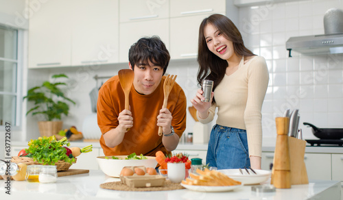 Asian young lovely lover couple husband and wife in casual outfit standing smiling holding red tomato while mixing vegetable salad with wooden fork and spoon in bowl in full decorated modern kitchen