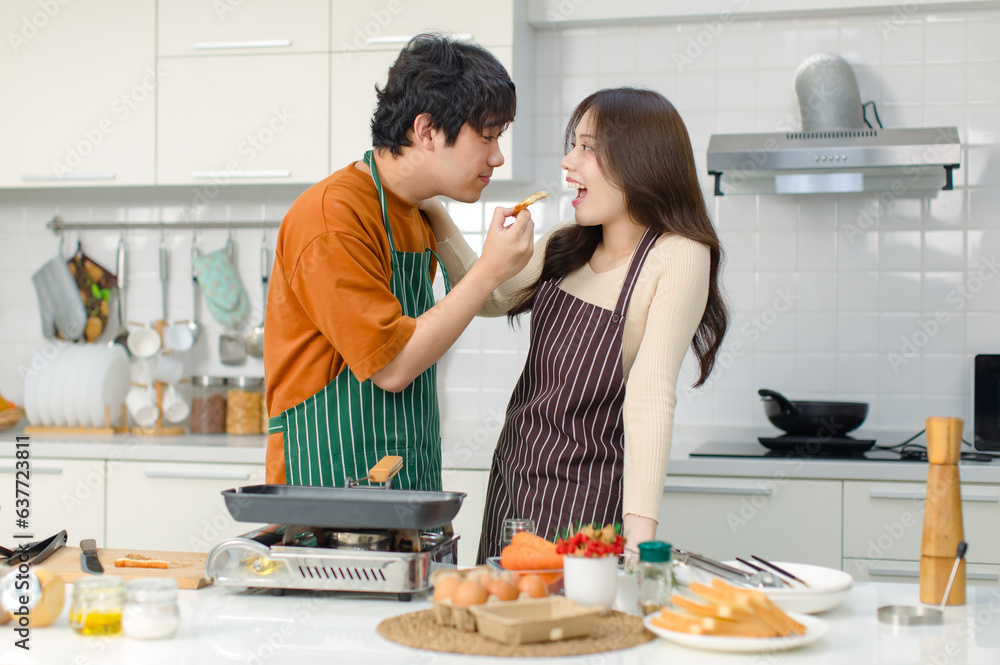 Asian young lover couple husband and wife in casual outfit with apron standing smiling holding frying half bread sandwiches posing together in full decorated modern kitchen with ingredients equipment