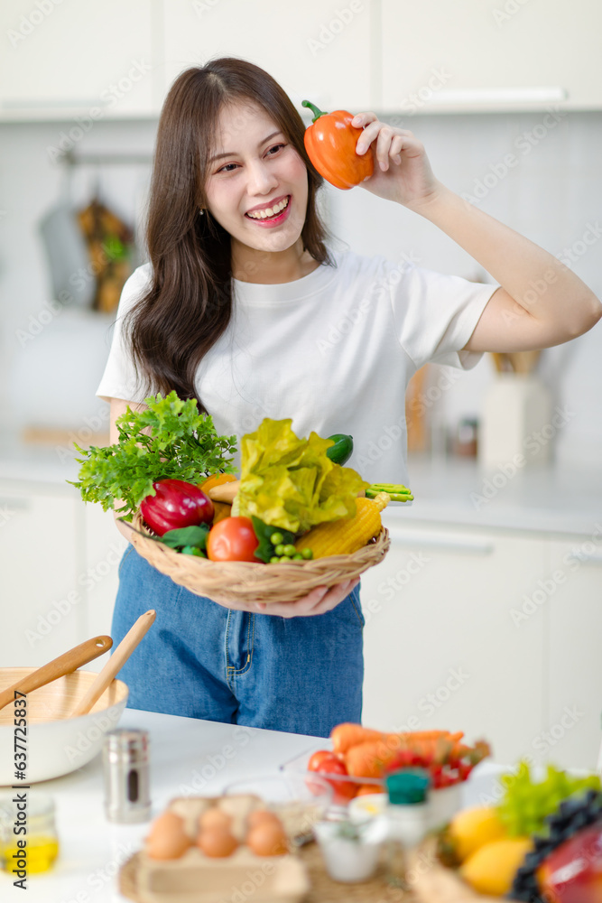 Asian young beautiful housewife in casual outfit standing smiling showing red sweet pepper and mixed fresh organic vegetables basket in full decorated modern kitchen with ingredients and equipment Asian young beautiful housewife in casual outfit standing smiling showing red sweet pepper and mixed fresh organic vegetables basket in full decorated modern kitchen with ingredients and equipment